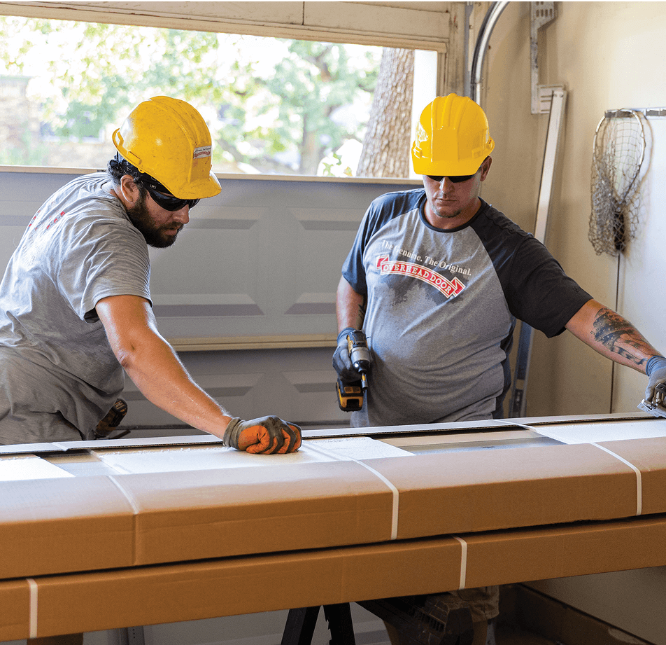 Two construction workers wearing yellow hard hats and gray shirts measure a large rectangular cardboard box in a garage. One worker holds a drill while the other adjusts the box with gloved hands.