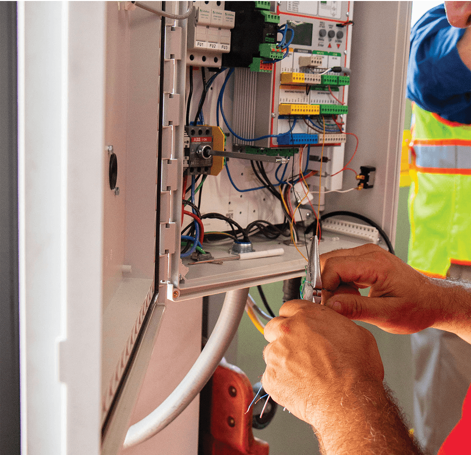 A person wearing a safety vest and blue shirt is working on electrical wiring inside an open control panel, using tools to connect wires. Various colored wires and electronic components are visible in the panel.