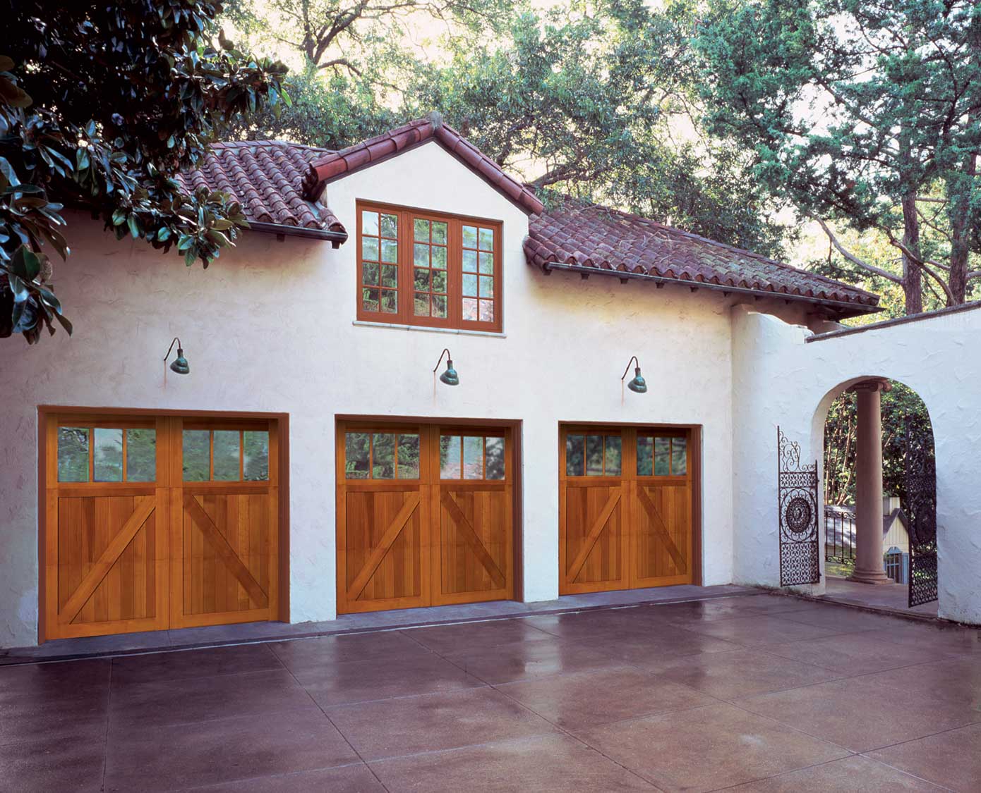 A stucco house with a red tile roof, three wooden garage doors with window panes, large driveway, and arched entryway sits surrounded by trees—perfect for those seeking trusted garage door installation services in Madison.