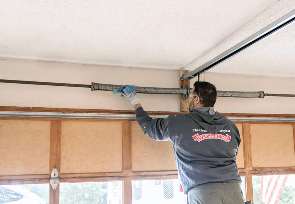 A man wearing gloves and a hoodie stands on a ladder inside a garage, performing service repair or inspecting a garage door torsion spring.