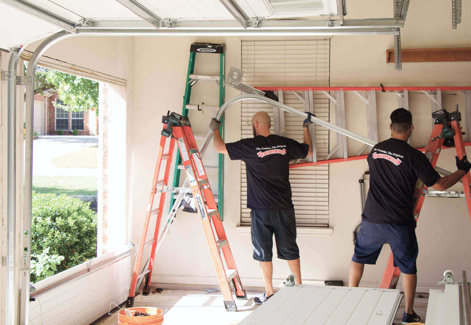 Two workers provide service repair on a garage door inside a sunlit garage, using ladders and tools. Both wear matching shirts with logos on the back as they work together to ensure the door functions smoothly.