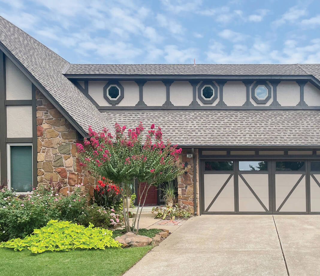 A Tudor-style house with stone and wood trim, octagonal windows, a double garage, and a blooming pink crepe myrtle tree in the landscaped front yard under a blue sky with clouds.