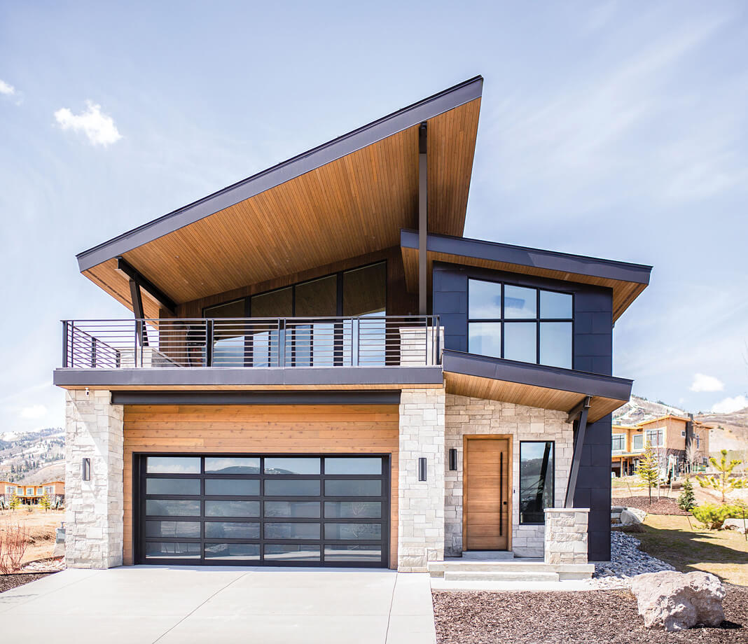 Modern house with a dramatic slanted roof, stone and wood accents, large windows, a glass-paneled garage door, and a spacious balcony above the garage. Blue sky and neighboring homes in the background.