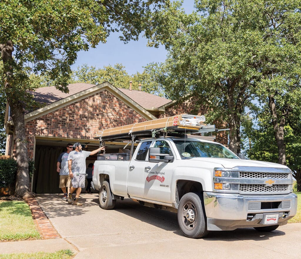 A white pickup truck labeled Roofing & Remodeling Services is parked in the driveway of a brick house. Two people are walking out of the open garage, carrying supplies. Trees and green lawn surround the home.