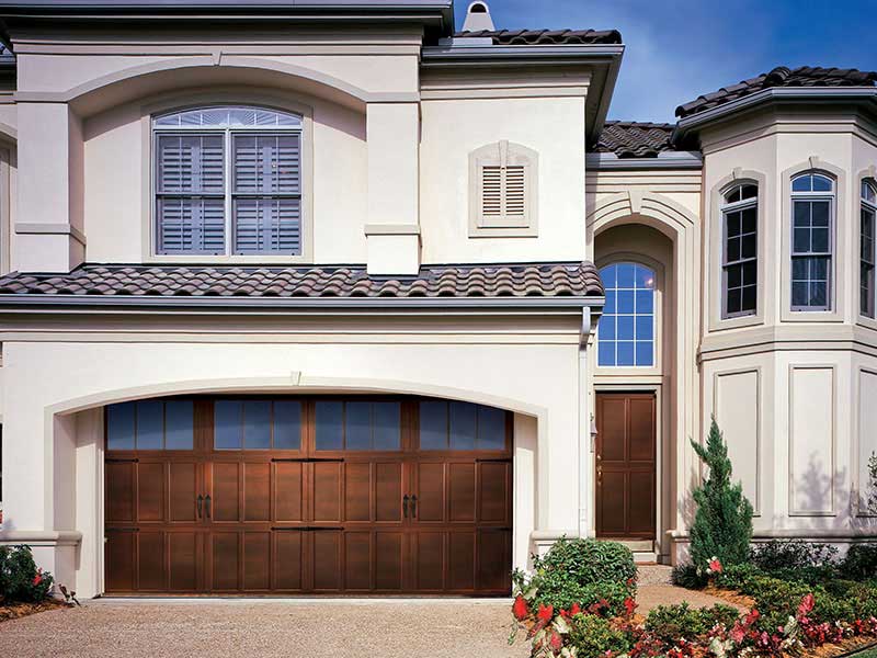A beige stucco house in Madison with a dark brown wooden garage door installed by trusted services, arched windows with white shutters, a side entry door, and landscaped bushes and flowers out front. The sky is clear and blue.