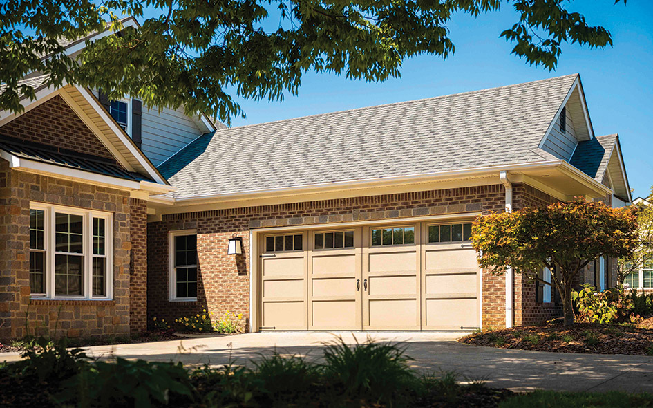 A suburban house with a brick exterior and two-car garage. The garage has cream-colored doors with windows at the top. Trees and landscaping surround the driveway under a clear blue sky.