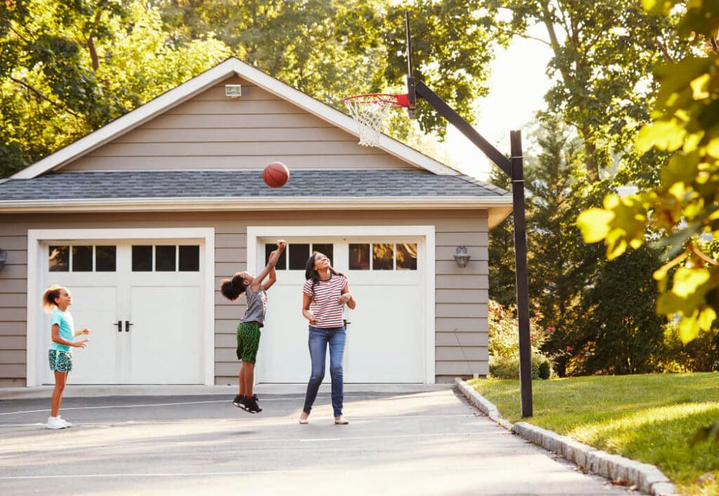 Three children play basketball in a driveway in front of a garage on a sunny day. One child shoots the ball toward the hoop while the others watch and stand nearby. Trees and greenery surround the area.
