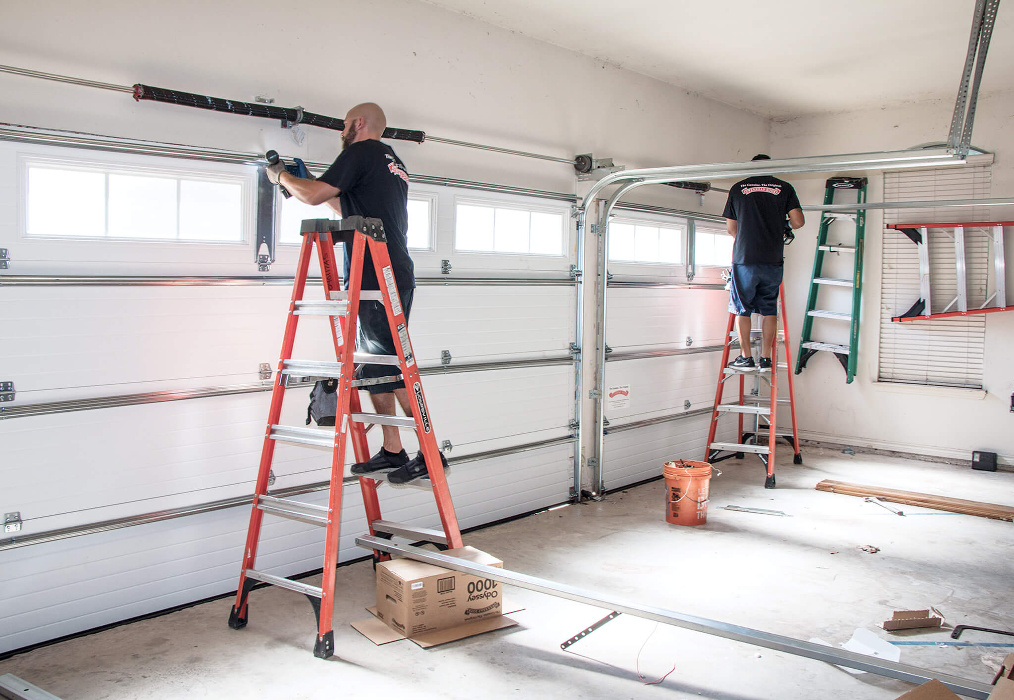 Two men standing on ladders perform a Service Repair on a large white garage door in a spacious, well-lit garage with scattered tools and materials on the floor.
