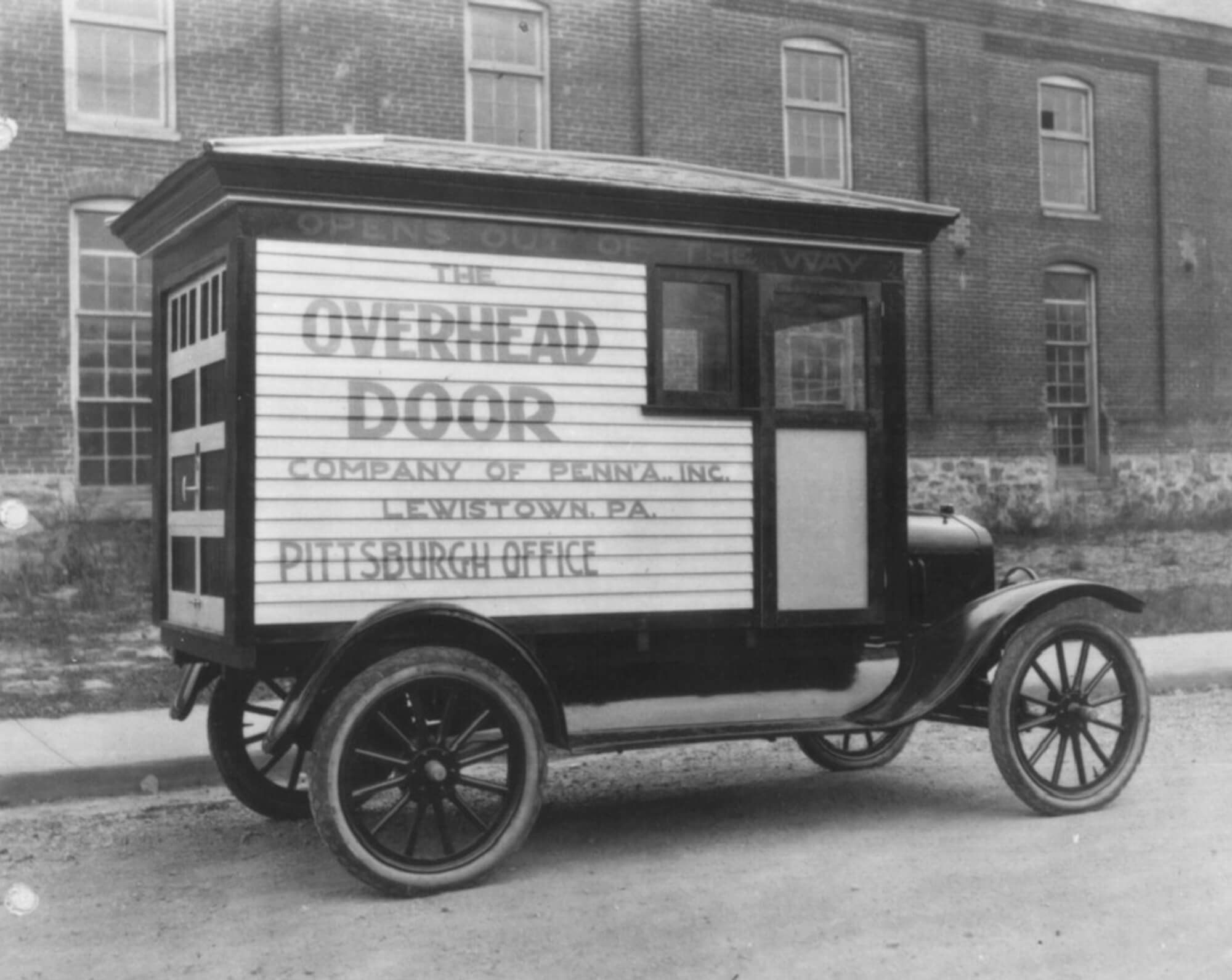A vintage delivery truck with The Overhead Door Company of Penna. Inc., Lewistown, PA, Pittsburgh Office—reflecting rich company background—parked on a street in front of a brick building.