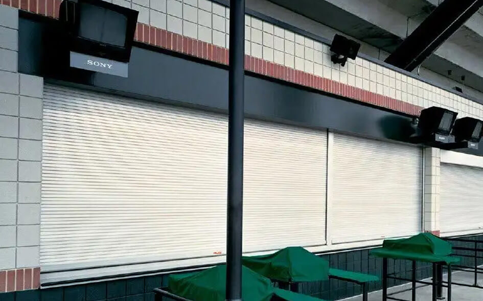 A row of metal rolling counter doors are closed over service windows at a building. Above the shutters, several mounted Sony TVs are visible, and in front stand green-covered tables. The scene appears empty and quiet.