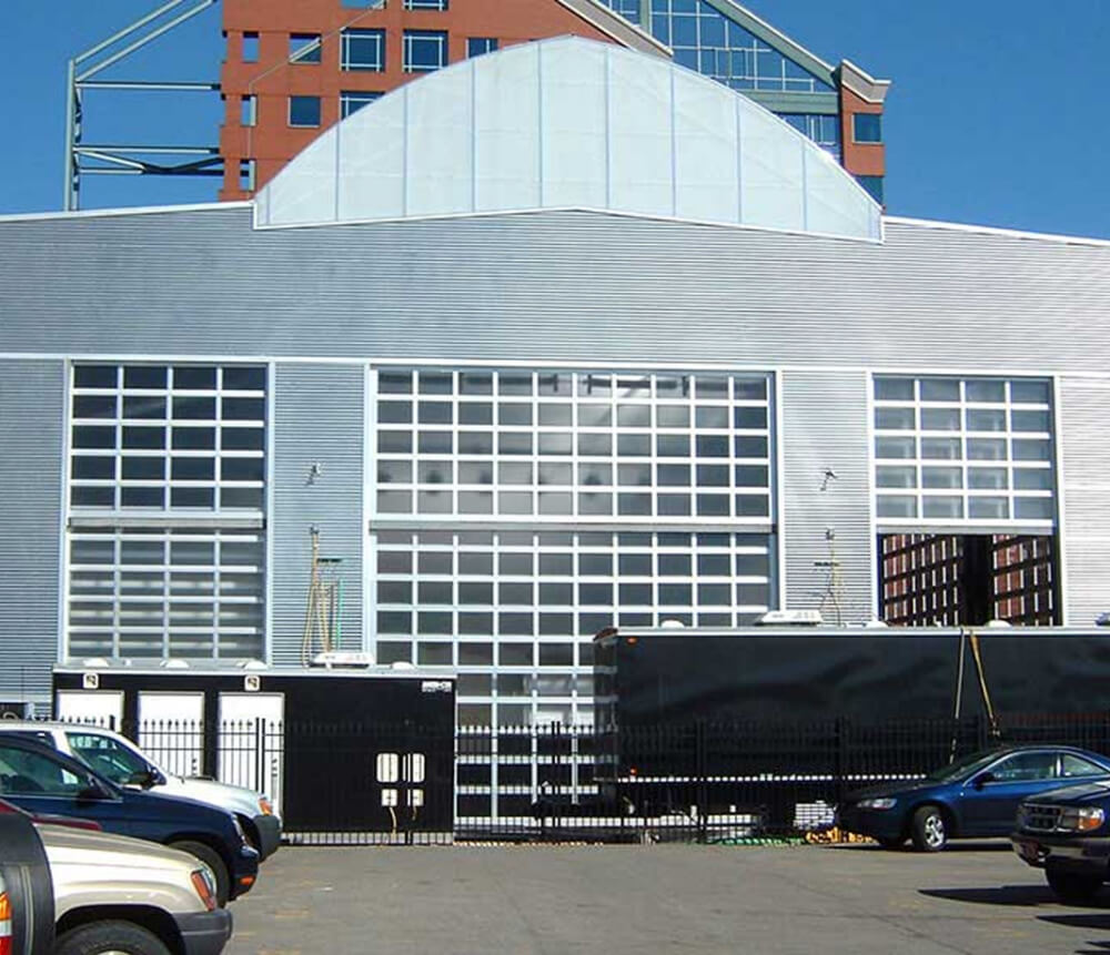 A large industrial-style building with metal siding, glass-paneled garage doors, and a geometric glass roof section. Cars are parked in front, and a black trailer is parked beside a black fence. A taller building is visible in the background.