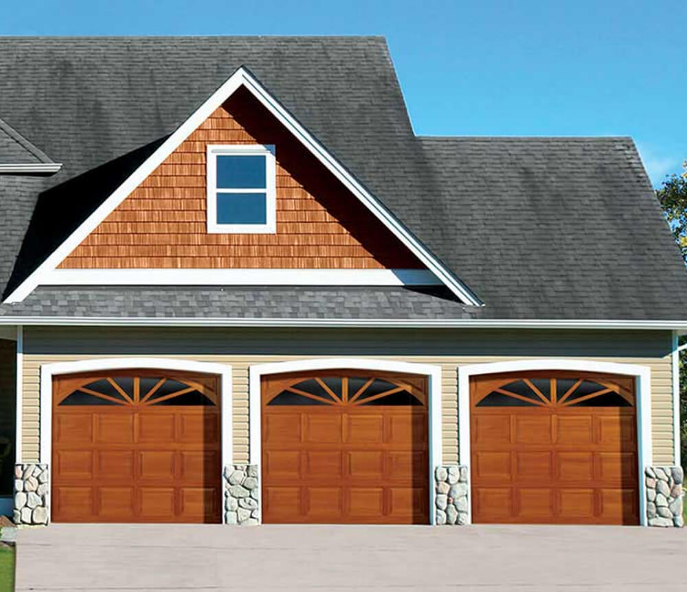 A house with three brown wooden garage doors, stone accents at the base of each door, yellow siding, and a steep gray shingled roof with a triangular gable and a single window above the garages.