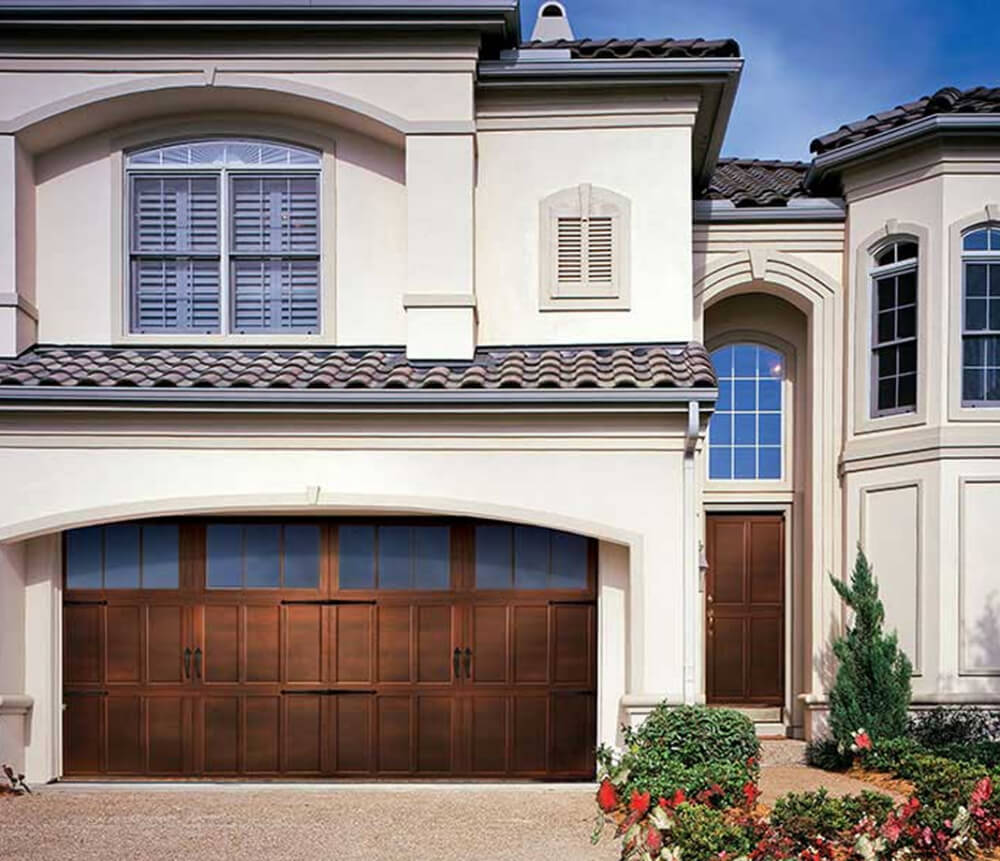 Large modern house with beige stucco walls, a brown double garage door with windows, arched windows with shutters, and a neatly landscaped front yard with flowers and greenery.