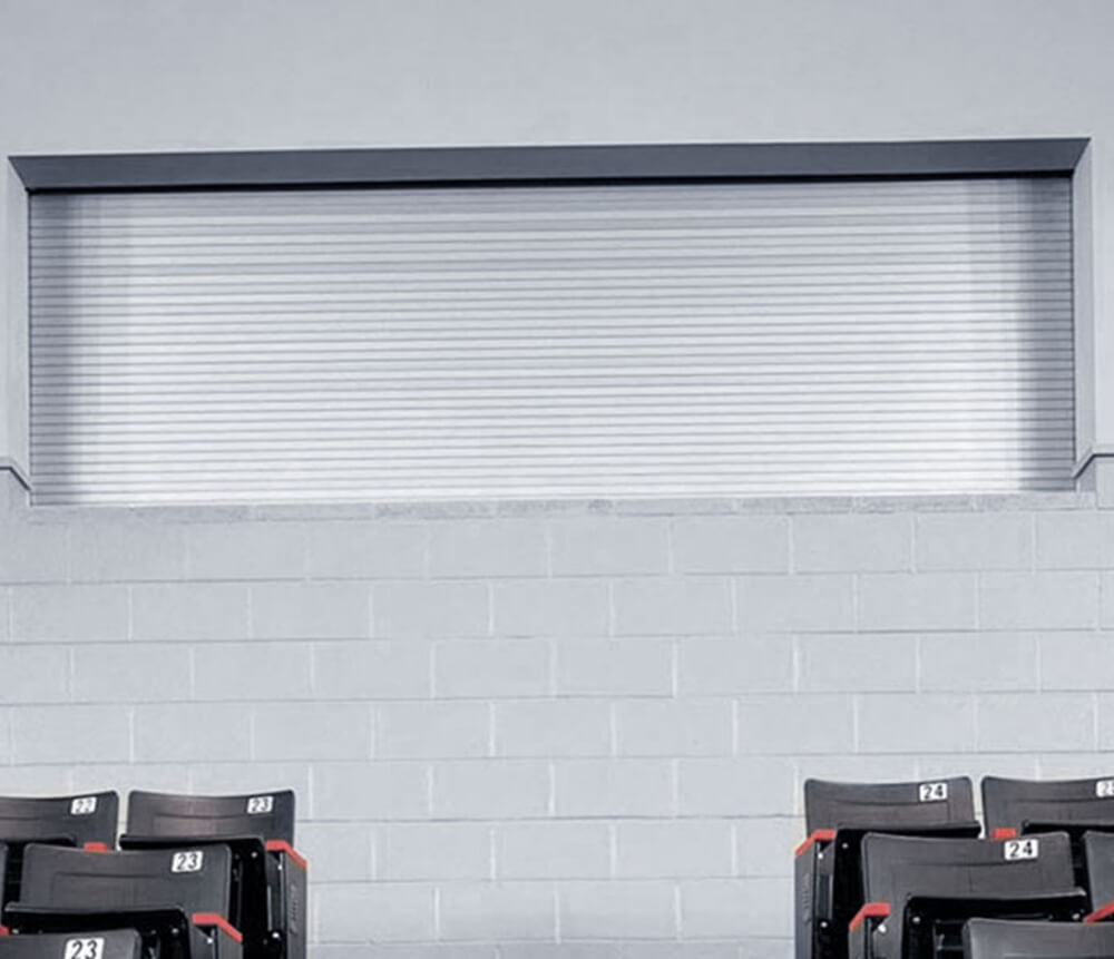 A closed metal rolling shutter is set in a gray cinder block wall above a row of empty black chairs with white number labels in a room with a minimalist, industrial look.