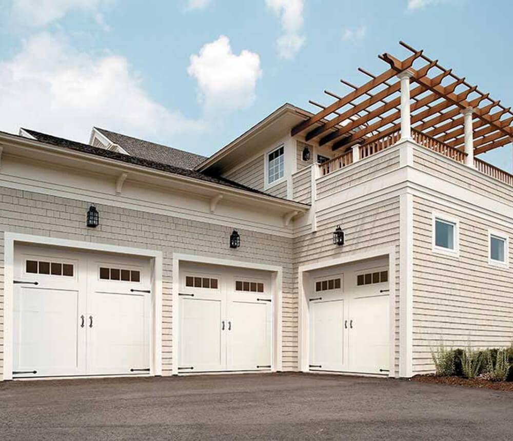 A beige house with three white garage doors, lantern-style lights, and a rooftop terrace featuring a wooden pergola. The driveway is paved, and the sky above is partly cloudy.