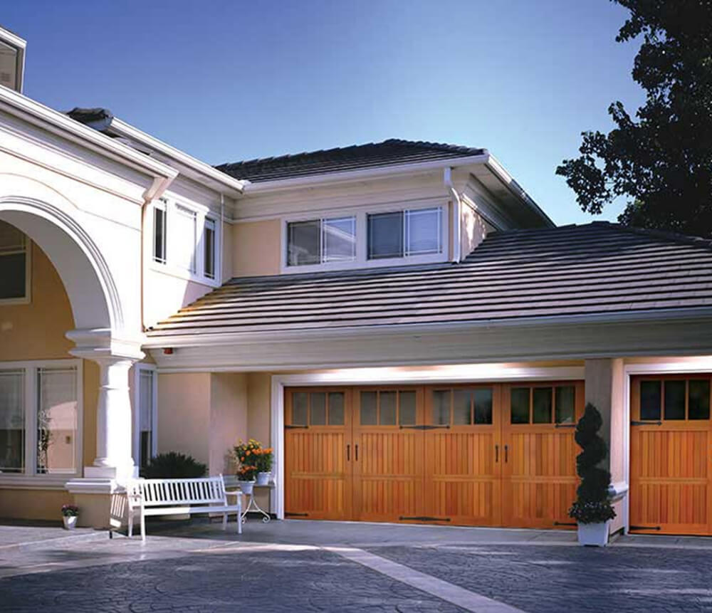 A modern house with a double wooden garage door, cream-colored exterior walls, a white bench near the entrance, and potted plants, under a clear blue sky.