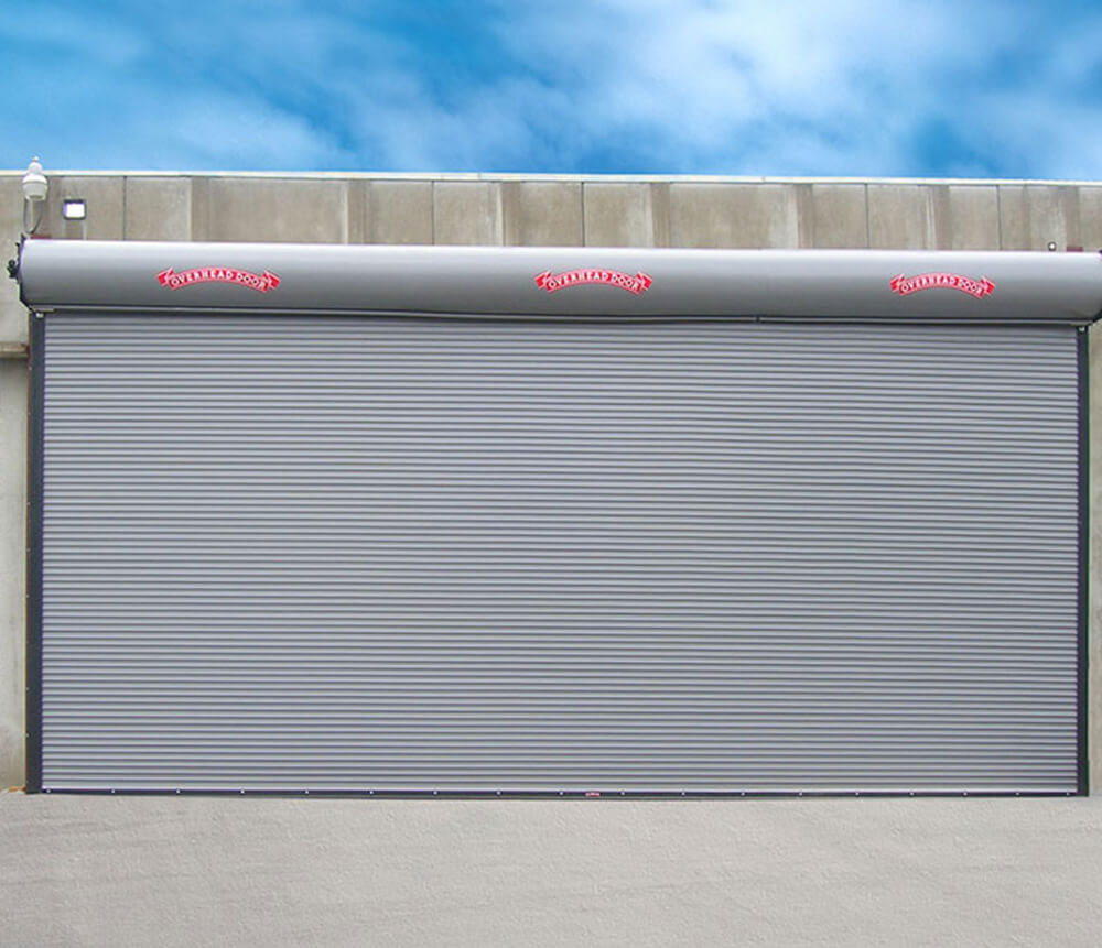 A large, gray industrial roller shutter door is closed on a concrete building under a blue sky with scattered clouds. The door has Bomber written in red at the top.