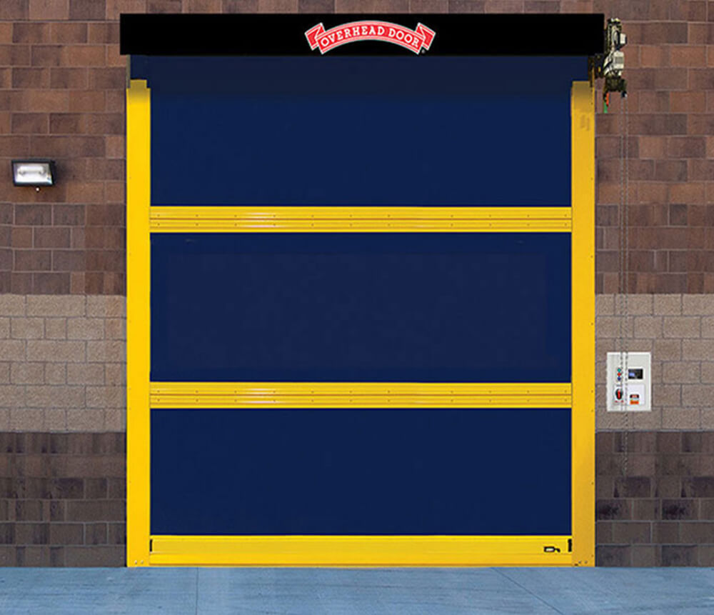 A blue industrial roll-up door with yellow framing is installed in a brick wall. A control panel is mounted on the right side, and a red sign above the door reads Overhead Door.