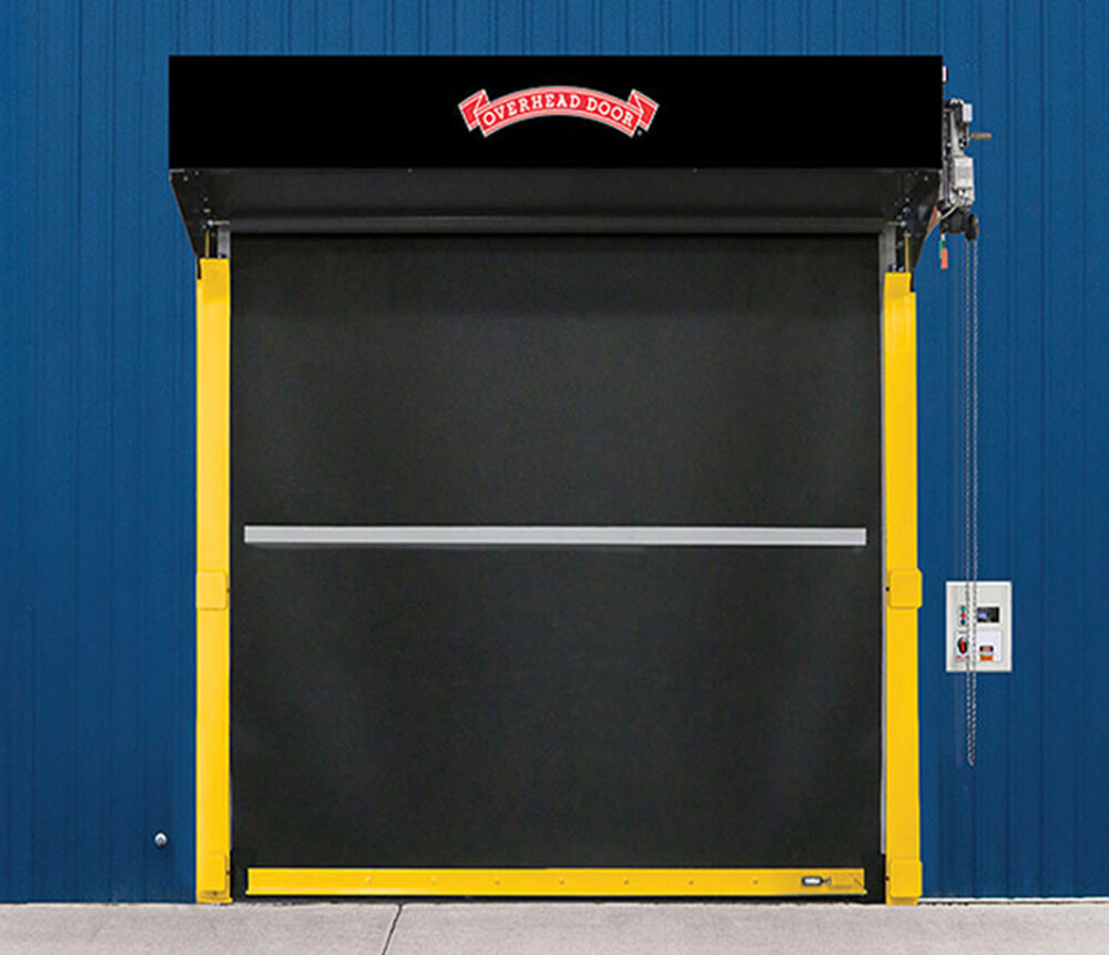 A black industrial roll-up door with a red Overhead Door logo is installed in a blue building. The door frame is yellow, and a control panel with a pull chain is mounted on the right side.