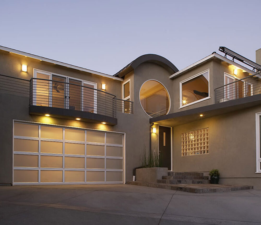 Modern two-story house with gray exterior, large windows, a circular window above the entrance, glass block wall, balcony, and illuminated garage and entryway lights at dusk.
