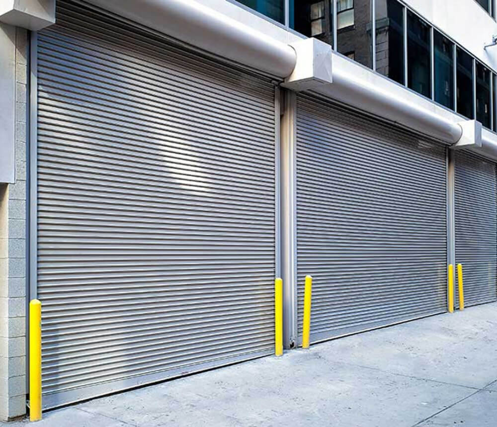 Three large silver metal roll-up security doors are closed on a building, with yellow bollards positioned in front of each door along the concrete sidewalk. The upper part of the building has glass windows.