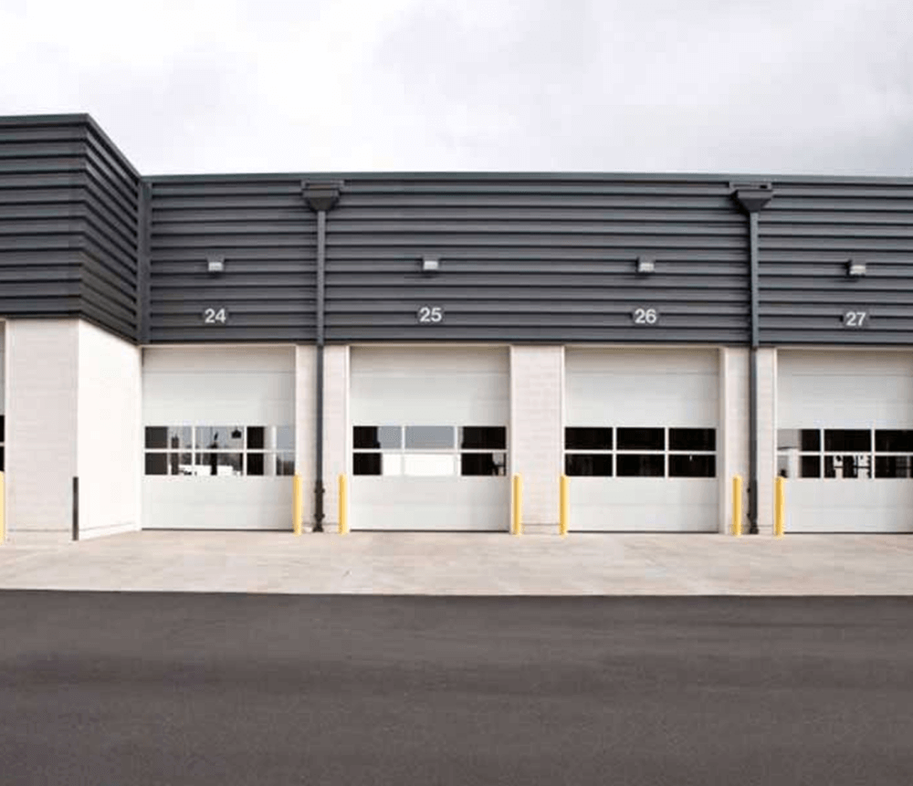 Modern industrial building exterior with four large garage doors numbered 24 to 27, yellow bollards in front, and an empty paved area under a cloudy sky.