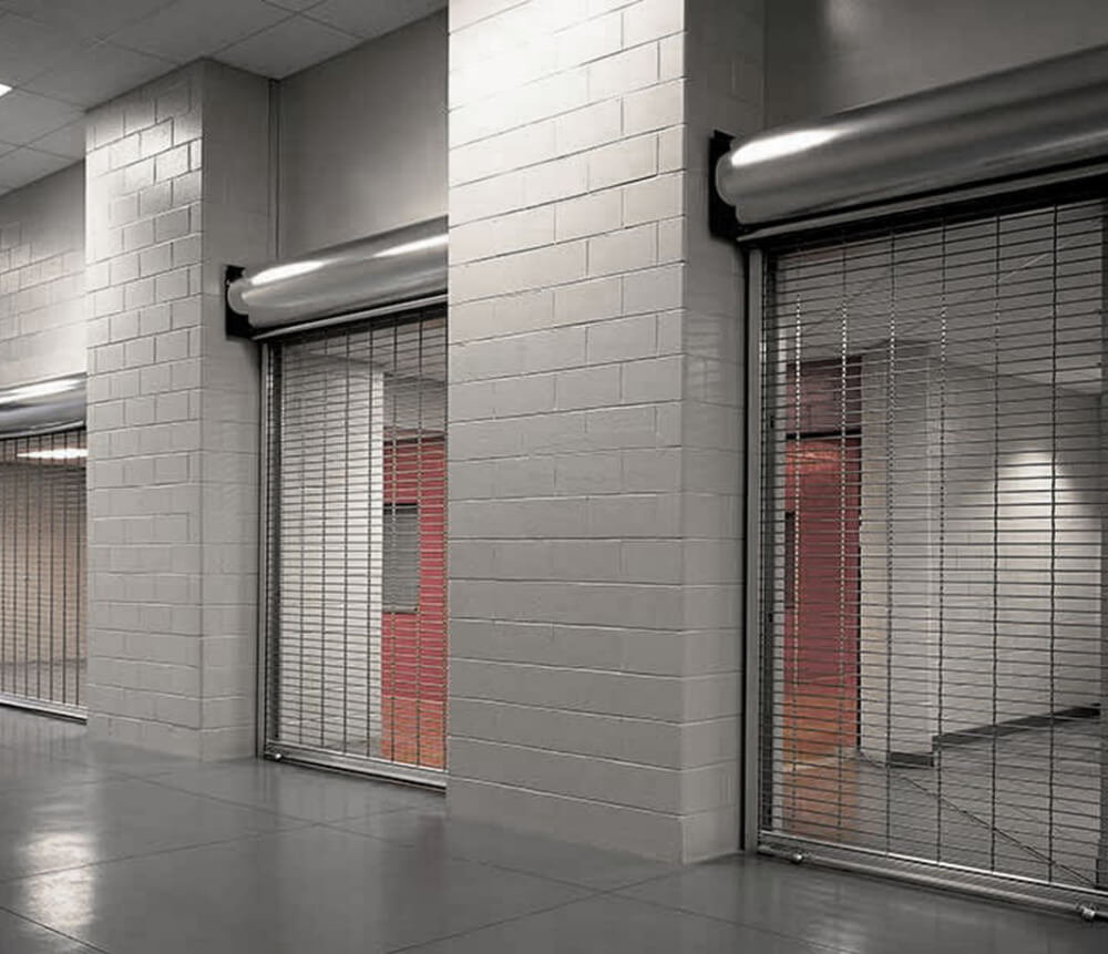 A hallway with large white tile walls and metal roll-up security grilles covering doorways, separating different sections of the building. The floor is smooth and gray.