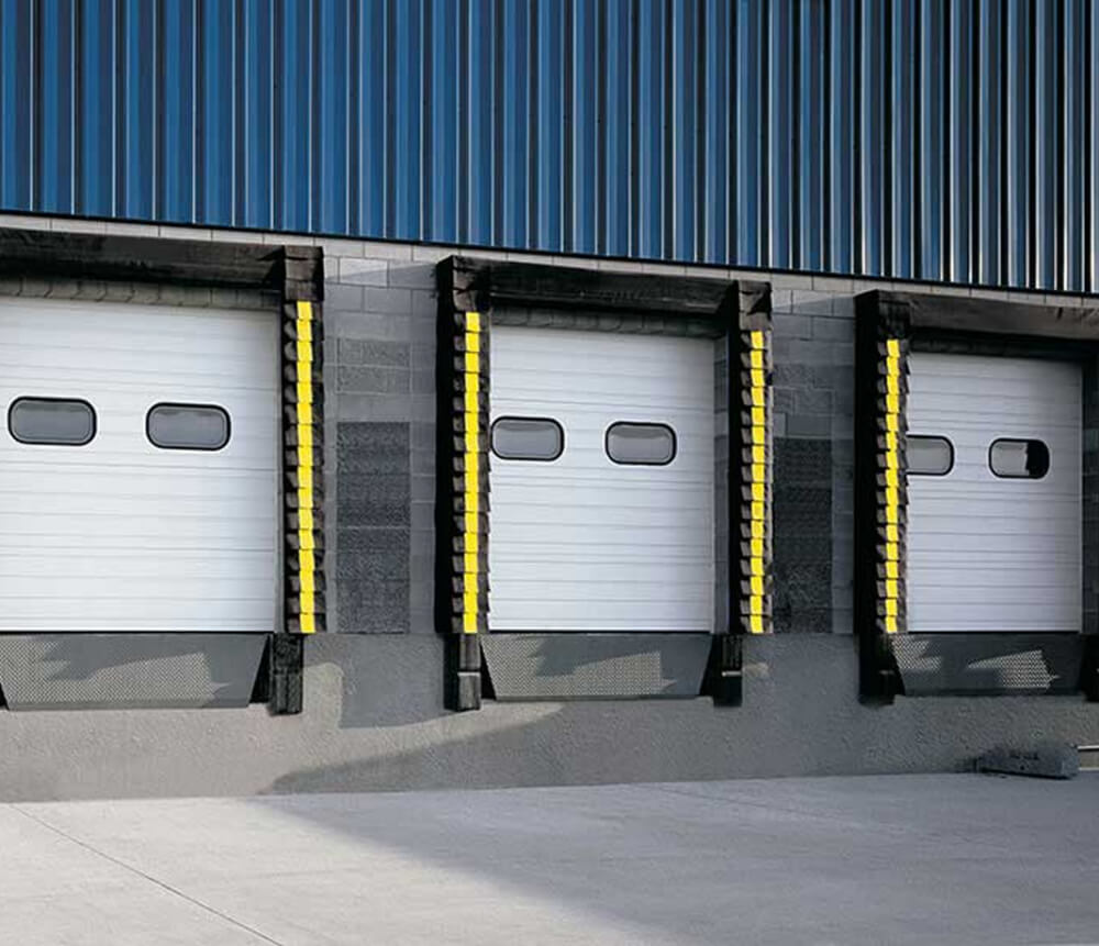 Three white industrial loading dock doors with small windows, surrounded by black and yellow protective bumpers, set in a gray and blue warehouse wall.