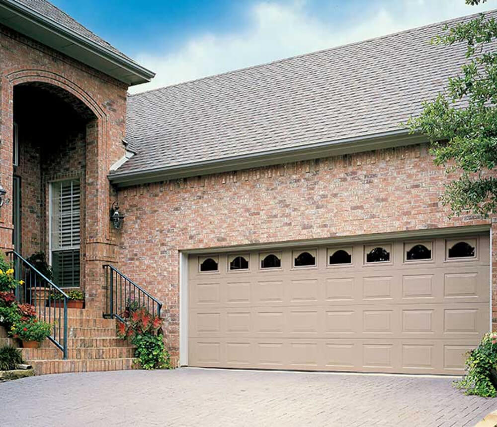 A brick house with a beige double garage door, small arched windows along the top, black metal railings by the steps, potted plants, and a clear, partly cloudy sky overhead.