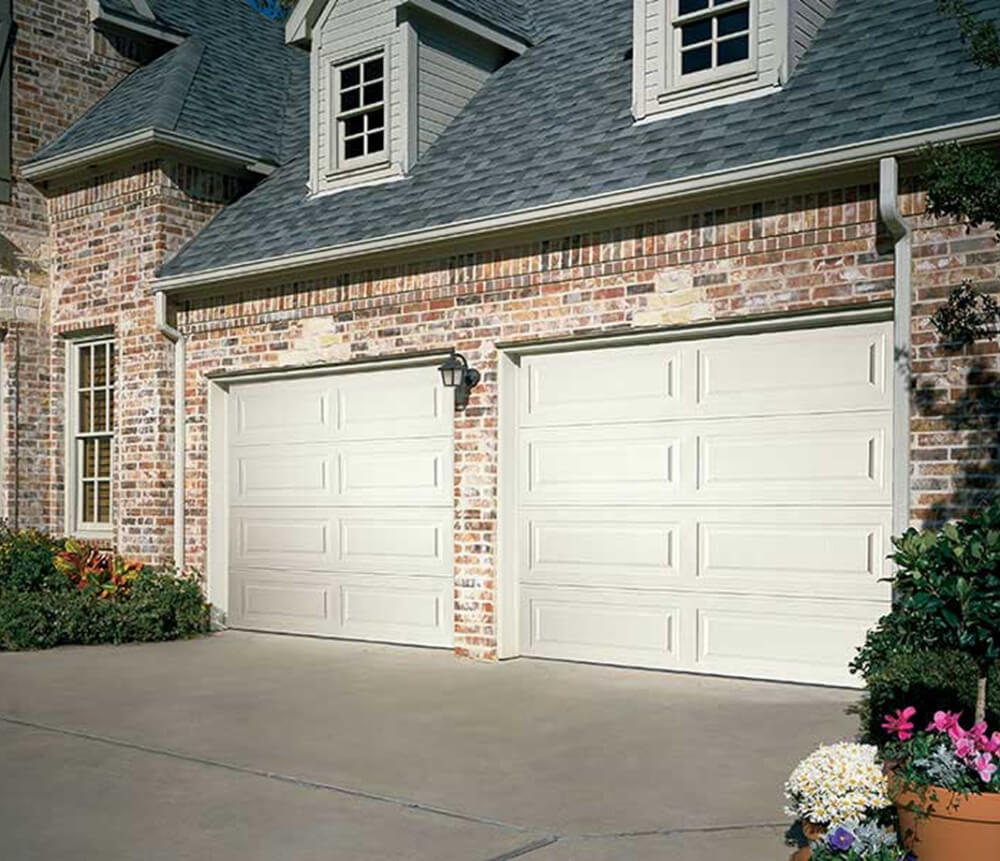 Two white, paneled garage doors on a brick house with gray roofing; a concrete driveway in front and a potted plant with colorful flowers in the lower right corner.