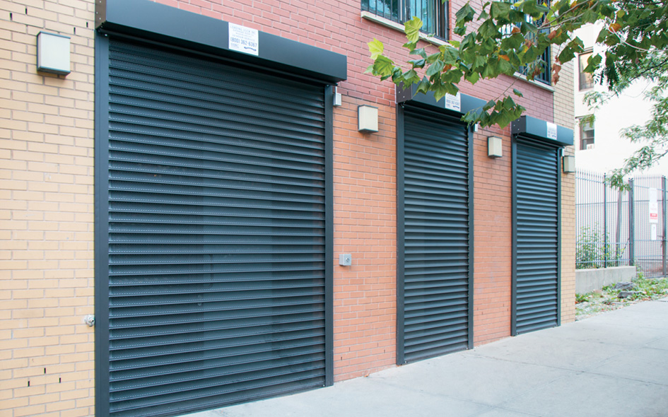 Three black metal Model 653 Allura roll-up garage doors set in a brick building, with a sidewalk and tree branches partially visible in the foreground.