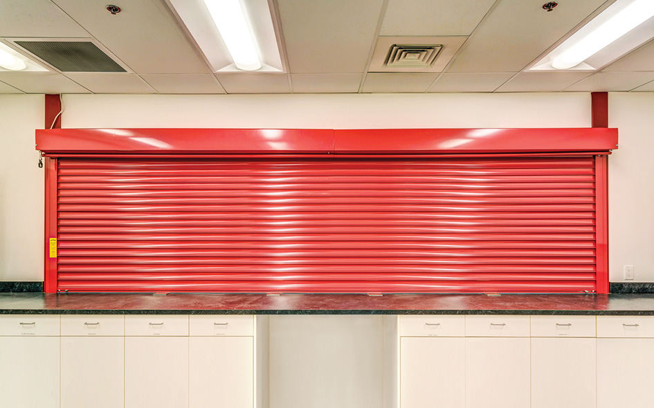 A bright red Allura Model 653 rolling shutter is closed over a wide service counter, with white cabinets below and fluorescent lights on the ceiling above.