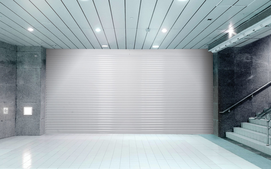 A closed silver Model 653 metal shutter door in a modern, empty building lobby with tiled flooring, gray stone walls, and a staircase with glass railings on the right, enhancing the sleek Allura-inspired design.