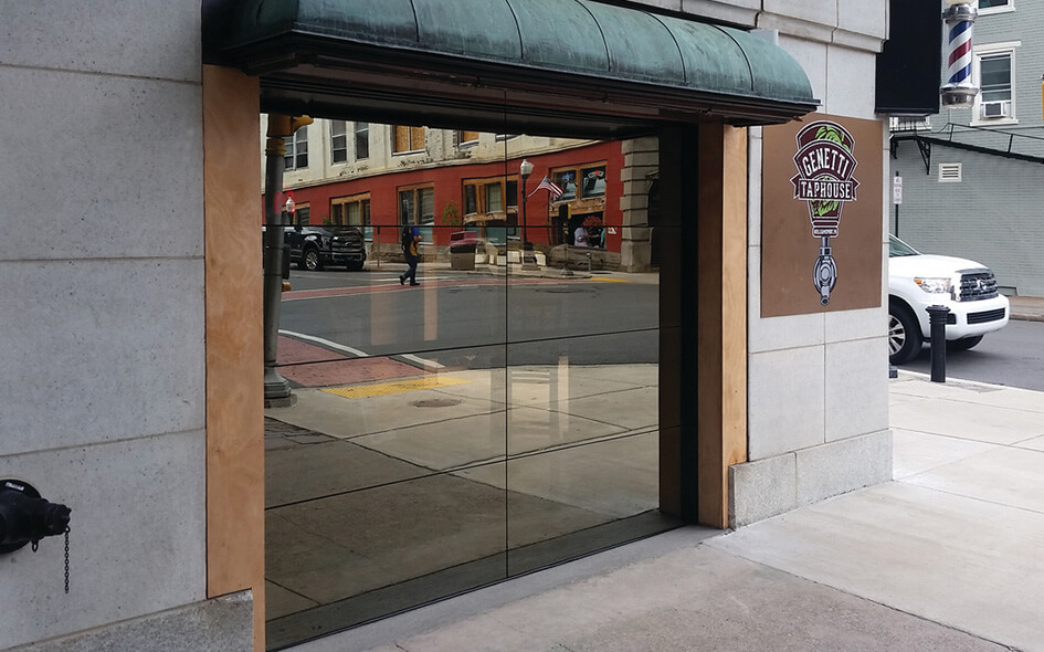 A Model 522 Aluminum Glass Door is partially open on a city street, reflecting cars and buildings. A sign reading Beauty & the Barber hangs on the wall next to the entrance.