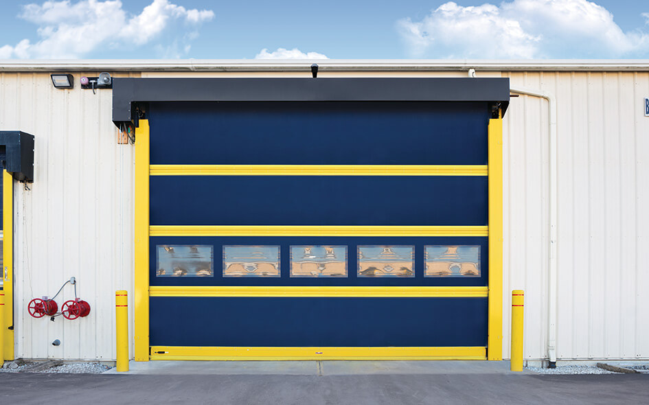 A large industrial overhead door with yellow and blue horizontal panels, set in a beige metal building. There are red valves on the left side of the wall and yellow bollards beside the door. The sky is partly cloudy.
