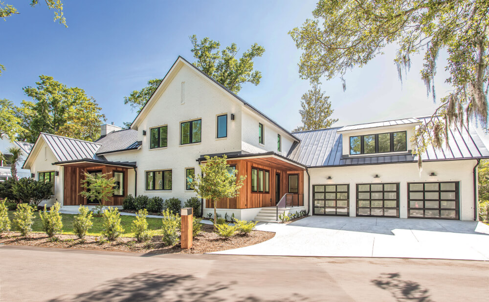 Modern white two-story house with black-framed windows, wooden accents, and a metal roof. There is a three-car garage with glass doors and a landscaped front yard with small trees and bushes under a clear blue sky.