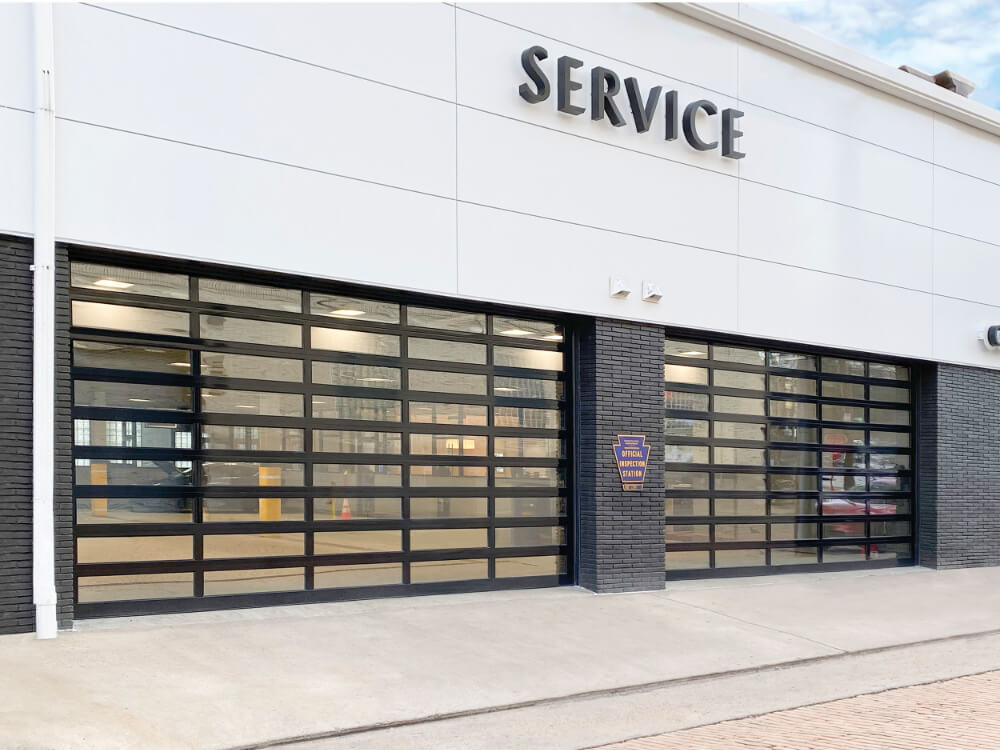 A modern building with large glass garage doors labeled SERVICE above the entrance, likely a service center or auto repair shop, with a paved sidewalk in front.