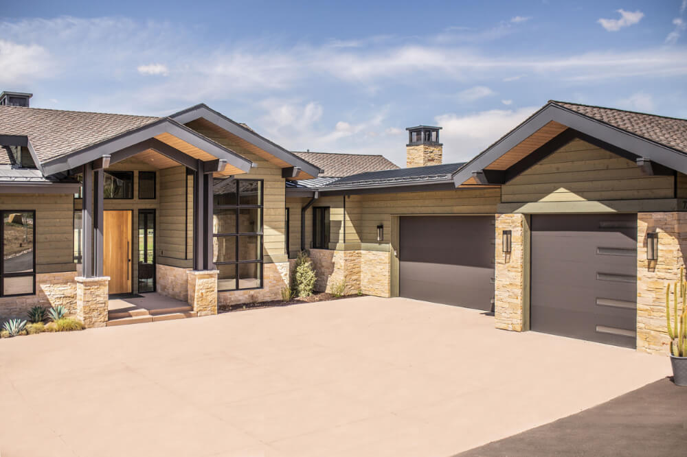 Modern house with stone and wood siding, large windows, two gray garage doors, and a spacious driveway under a partly cloudy sky.