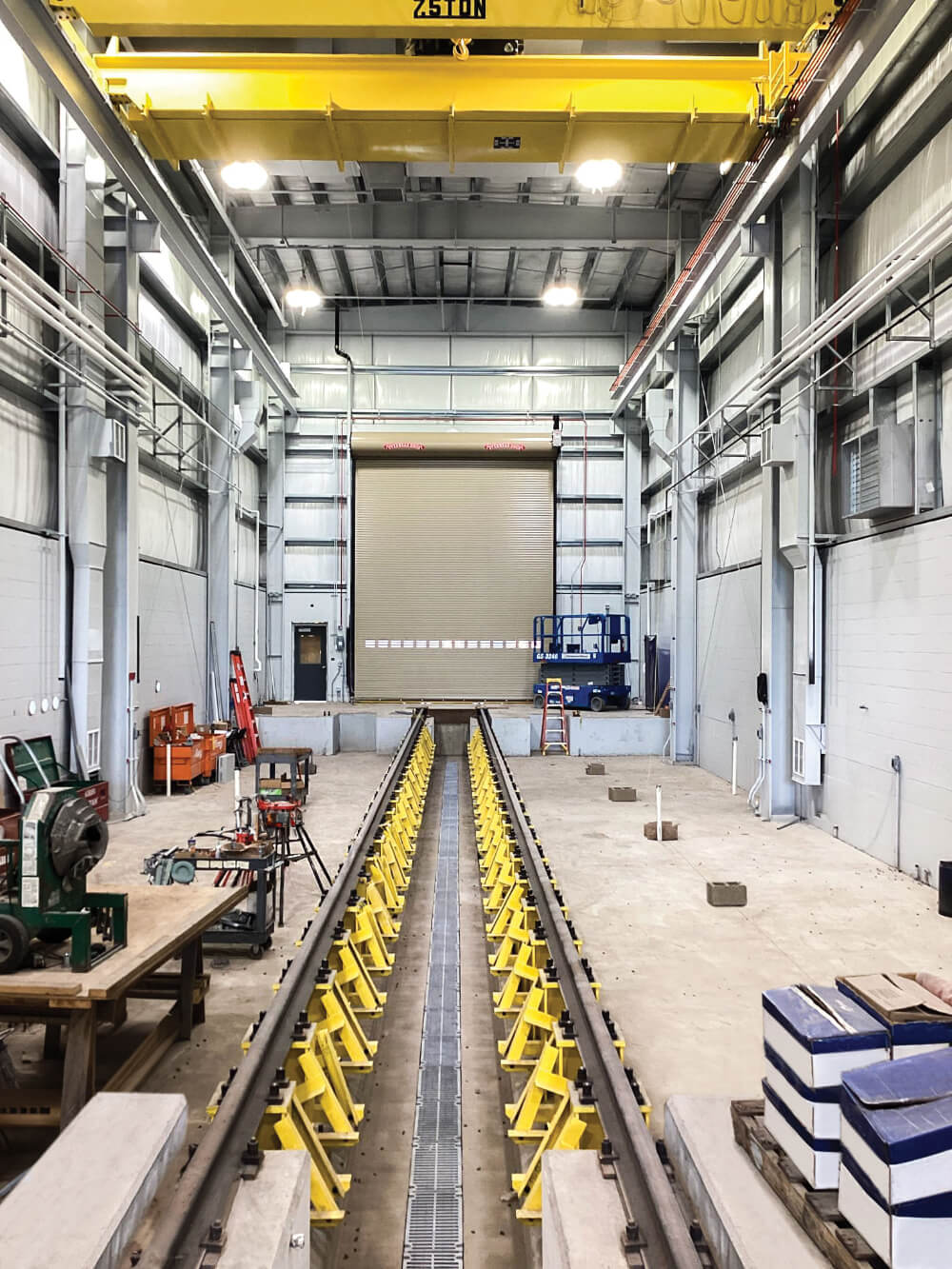 Interior of an industrial facility with metal tracks running down the center, surrounded by machinery, tools, and workbenches. A large yellow crane is mounted overhead, and the space is brightly lit with high ceilings.