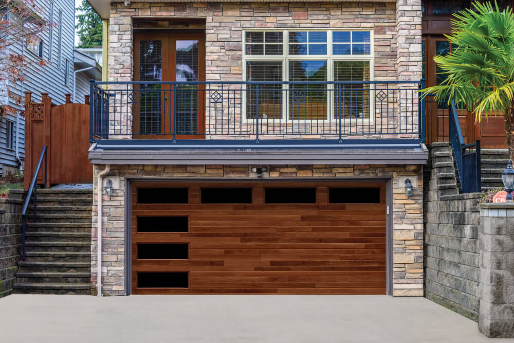 A modern house exterior with stone walls, a wooden garage door featuring rectangular windows, a balcony with a metal railing, and stairs on either side leading to the entrance.