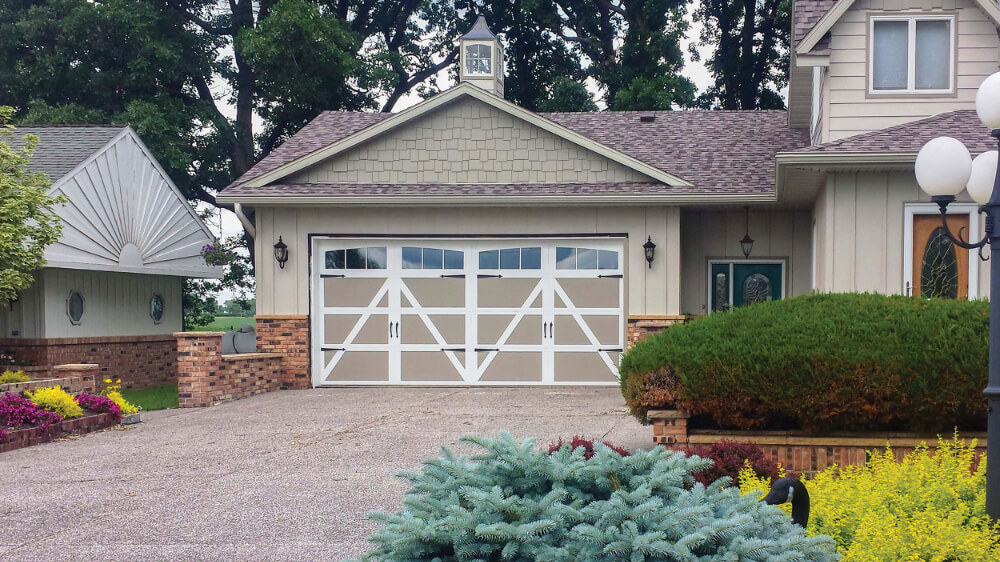A suburban house with a decorative double garage door, surrounded by landscaped bushes, colorful flowers, and trimmed shrubs. The driveway is paved, and the house features tan siding with brick accents.