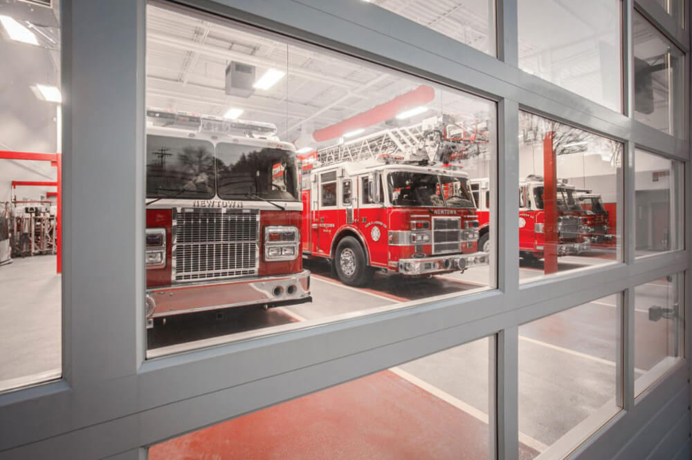 View through large windows of a fire station showing several red fire trucks parked in a well-lit garage, with equipment and tools visible in the background.