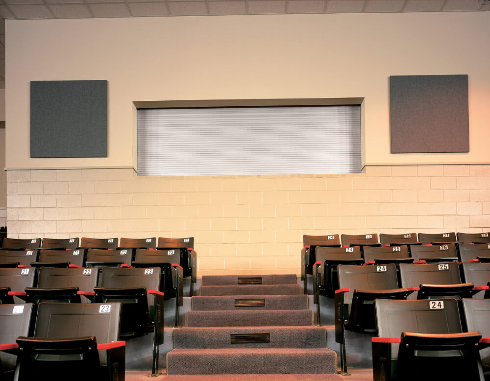 Rows of empty black auditorium seats with seat numbers, red armrests, and a central carpeted staircase face a cream wall featuring Door 651—a closed rolling counter door—and two gray acoustic panels.