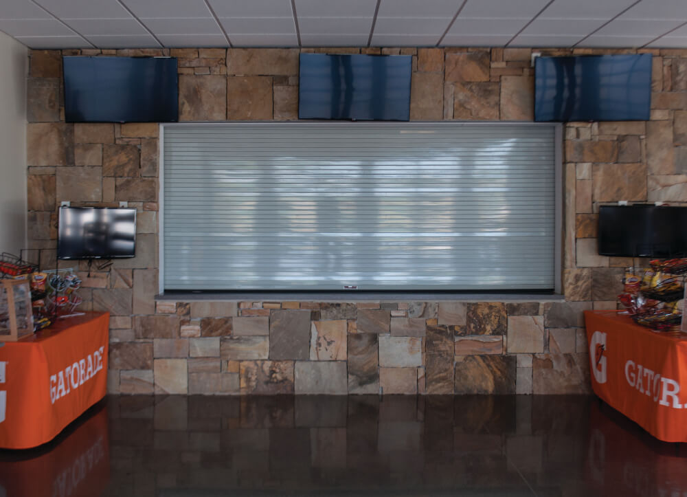 A closed concession stand with a metal rolling counter door, surrounded by stone walls. Four TV screens are mounted above, and tables with orange Gatorade tablecloths and snacks are set up on either side.