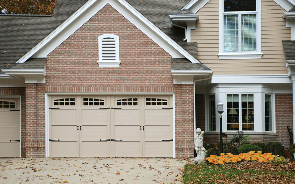 A suburban house with a double beige garage door, red brick exterior, white trim, a large window, and a small statue near a garden bed with yellow flowers.