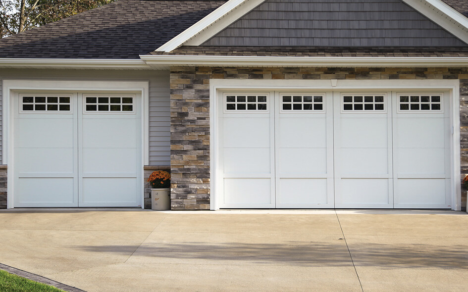 Two white garage doors with small square windows at the top are set in a house exterior with gray siding and stone accents; a potted plant with orange flowers sits beside the left door on a clean driveway.