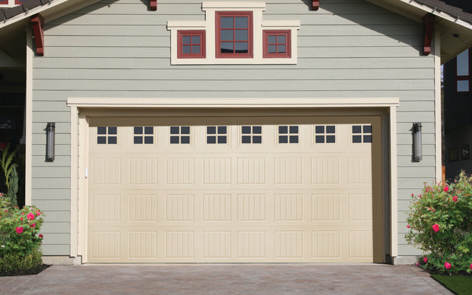 A beige double garage door from the Durafirm Collection features rectangular windows along the top panel, set in a light green house with white trim, red window frames, and small bushes with pink flowers flanking the driveway.