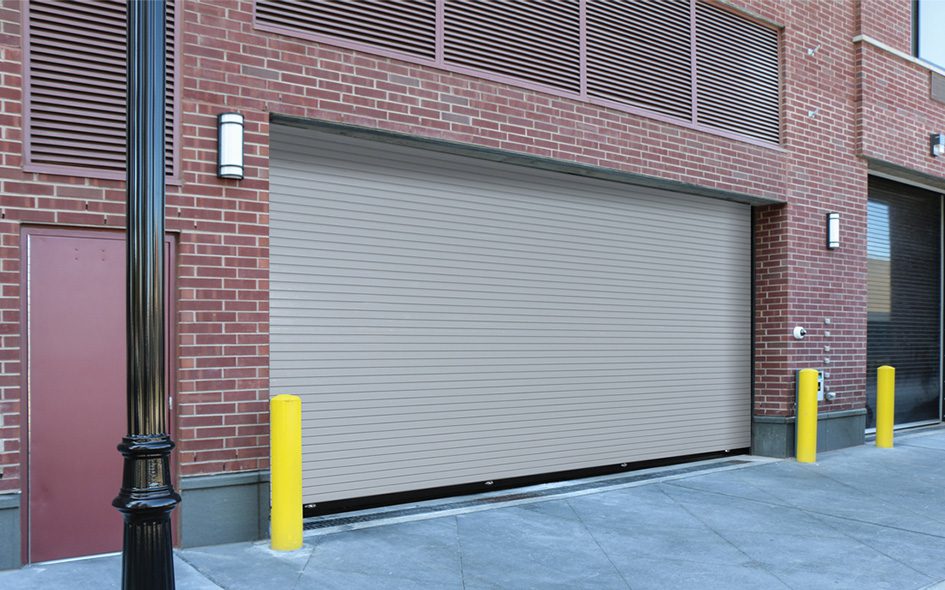 A beige 610S Springless Service Door in a brick building, flanked by four yellow safety bollards and a black streetlamp, with a red access door and vent panels on the left.