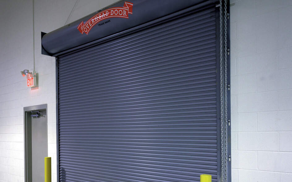 A large industrial gray rolling service door (631) is closed in a warehouse setting. The door, possibly a fire-rated door, has a red Overhead Door sign above it and is flanked by yellow safety bollards, with an emergency exit nearby.