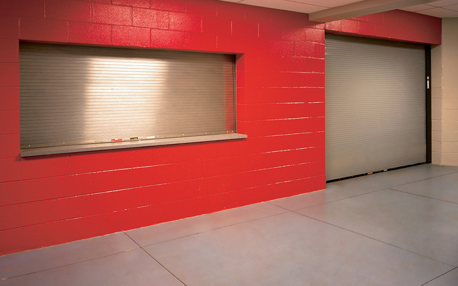 A red-painted wall features a closed metal concession window with a fire rated rolling door and a large closed rolling door, all set in an indoor space with a light blue floor and beige ceiling tiles.